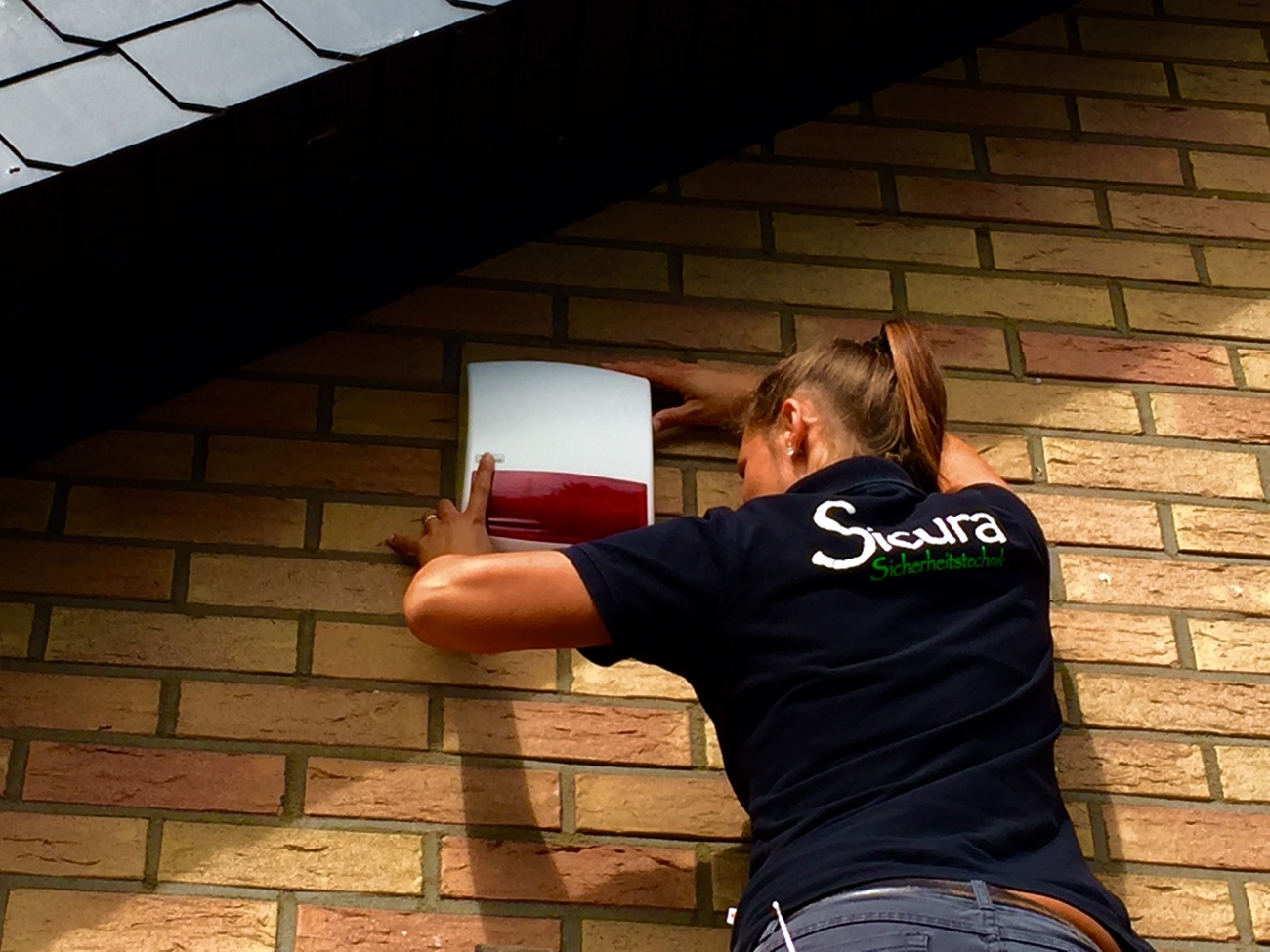 Sicura-Technikerin montiert eine Außensirene an einer Klinkerfassade